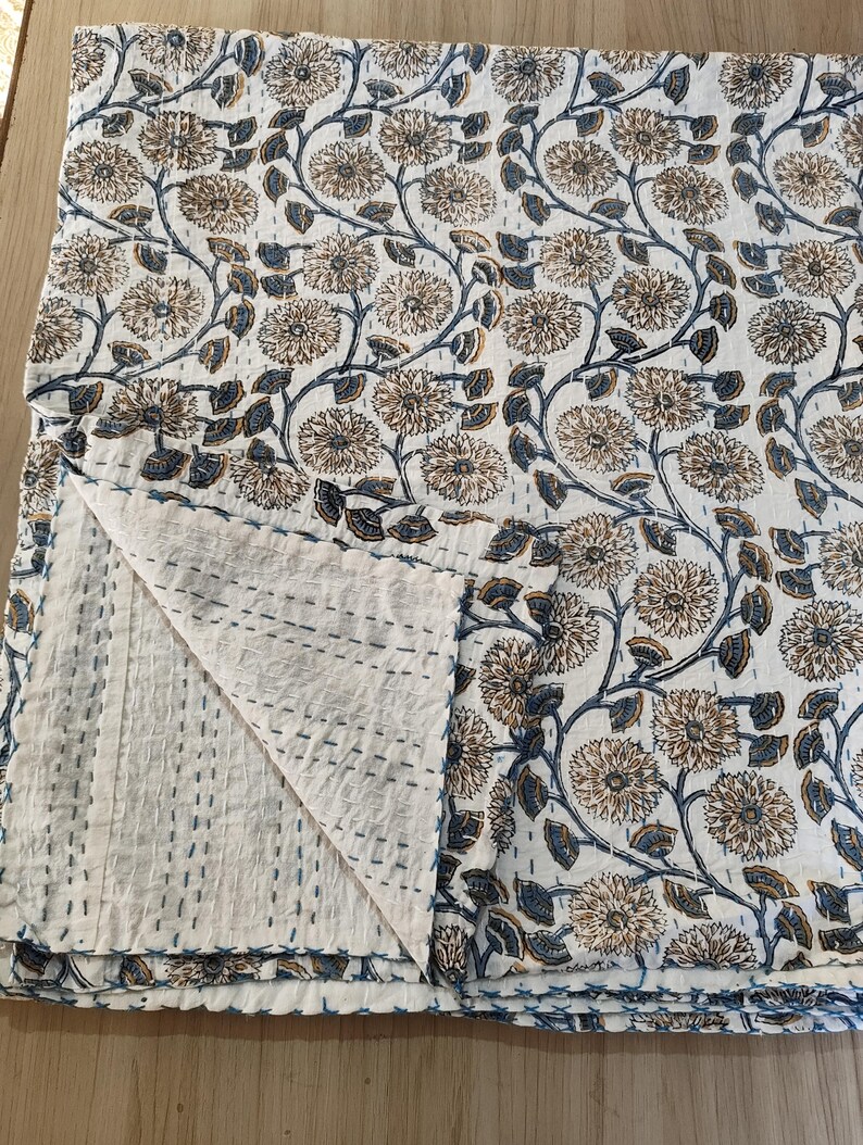K&ouml;nnte beinhalten: Eine gefaltete wei&szlig;e Steppdecke mit einem floralen Muster in Braun- und Blaut&ouml;nen. Die Steppdecke weist ein sich wiederholendes Design aus stilisierten Blumen und Ranken auf. Die Kanten sind mit blauen N&auml;hten versehen, und die Steppdecke ist gefaltet, um die Unterseite freizulegen.