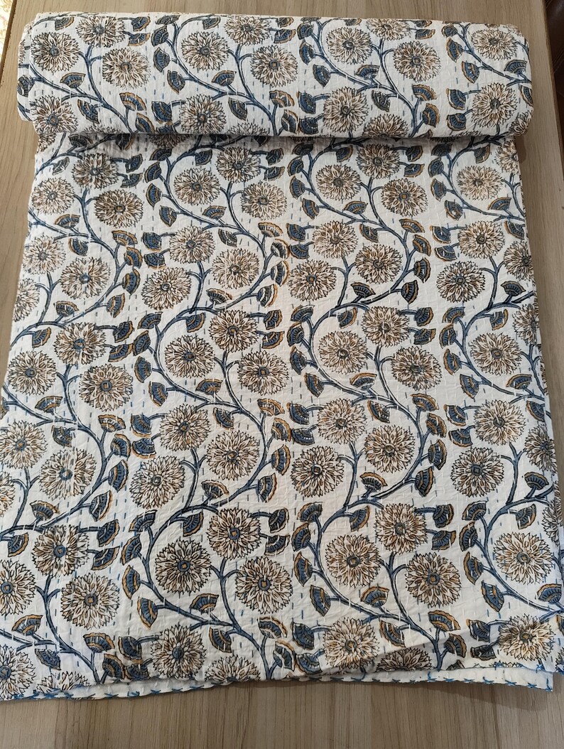 K&ouml;nnte beinhalten: Eine aufgerollte Steppdecke mit floralem Muster. Die Steppdecke zeigt ein sich wiederholendes Muster aus gro&szlig;en, hellbraunen Blumen mit blauen Stielen und Bl&auml;ttern auf wei&szlig;em Hintergrund. Die Steppdecke besteht aus einem weichen, gesteppten Stoff.