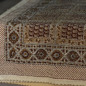May include: A detailed close-up of a brown and cream-colored textile with an intricate floral and geometric pattern. The edges feature a striped design and fringe. The textile is likely a decorative item, such as a throw or tablecloth.