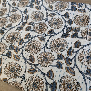 K&ouml;nnte beinhalten: Ein wei&szlig;es Textil mit einem sich wiederholenden Blumenmuster. Das Design zeigt gro&szlig;e, stilisierte Blumen in Braun- und Beiget&ouml;nen mit blau-grauen Bl&auml;ttern und Stielen. Der Stoff scheint gesteppt zu sein.