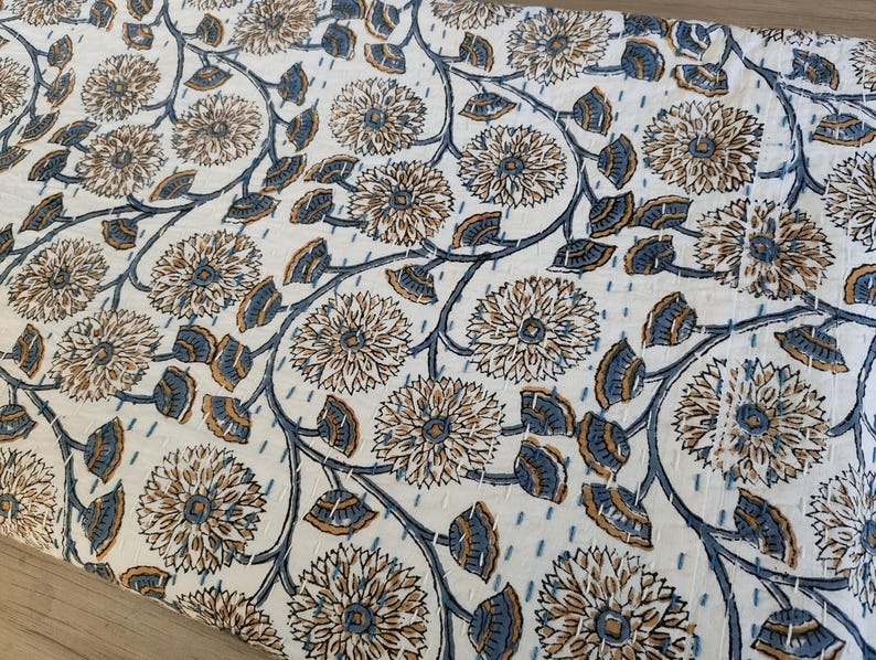 K&ouml;nnte beinhalten: Ein wei&szlig;es Textil mit einem sich wiederholenden Blumenmuster. Das Design zeigt gro&szlig;e, stilisierte Blumen in Braun- und Beiget&ouml;nen mit blau-grauen Bl&auml;ttern und Stielen. Der Stoff scheint gesteppt zu sein.