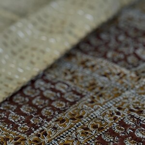 May include: Close-up of a folded textile with a detailed pattern. The fabric features a combination of brown, tan, and white colors. The design includes floral and geometric elements, creating a textured appearance. The stitching is visible, adding to the handcrafted look.