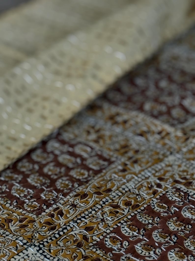 May include: Close-up of a folded textile with a detailed pattern. The fabric features a combination of brown, tan, and white colors. The design includes floral and geometric elements, creating a textured appearance. The stitching is visible, adding to the handcrafted look.