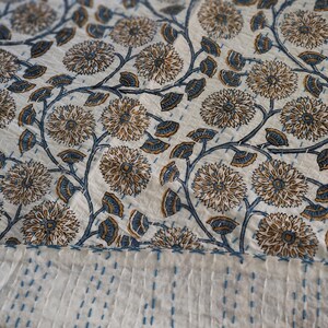 K&ouml;nnte beinhalten: Ein wei&szlig;es Textil mit einem Blumenmuster in Braun- und Blaut&ouml;nen. Das Design zeigt gro&szlig;e, stilisierte Blumen und Bl&auml;tter auf sich windenden Stielen. Der untere Rand hat einen Abschnitt mit blauen N&auml;hten, was auf eine Steppdecke oder einen &auml;hnlichen Artikel hindeutet.