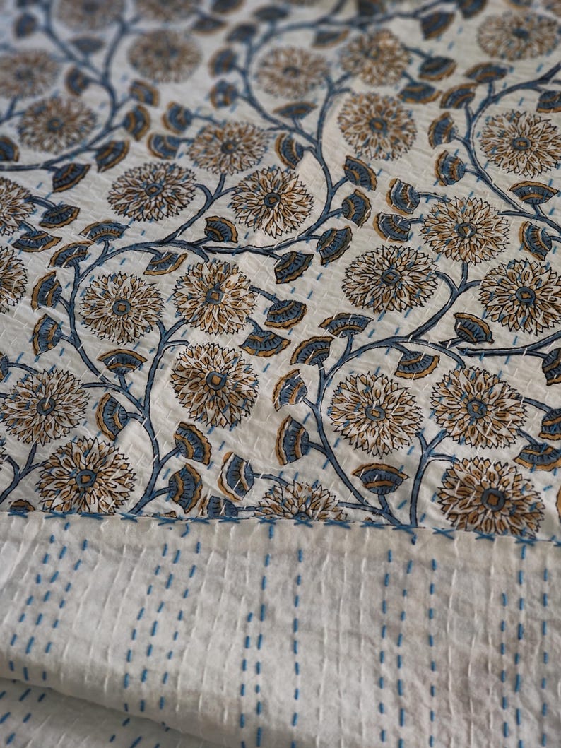 K&ouml;nnte beinhalten: Ein wei&szlig;es Textil mit einem Blumenmuster in Braun- und Blaut&ouml;nen. Das Design zeigt gro&szlig;e, stilisierte Blumen und Bl&auml;tter auf sich windenden Stielen. Der untere Rand hat einen Abschnitt mit blauen N&auml;hten, was auf eine Steppdecke oder einen &auml;hnlichen Artikel hindeutet.