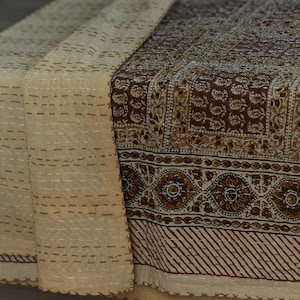 May include: A cream-colored textile with a brown floral pattern and decorative border. The fabric is stitched with white thread, and the edges are scalloped. The textile is folded over, revealing the underside with a cream color and stitched details.