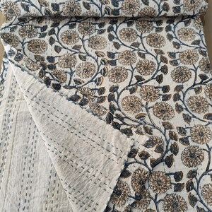 K&ouml;nnte beinhalten: Eine wei&szlig;e und beige gesteppte Decke mit floralem Muster und blauen Akzenten. Die Steppdecke hat ein sich wiederholendes Muster aus gro&szlig;en Blumen und Bl&auml;ttern. Die Kanten sind mit blauem Faden gen&auml;ht. Die Steppdecke ist gefaltet und aufgerollt, wodurch das Muster sichtbar wird.