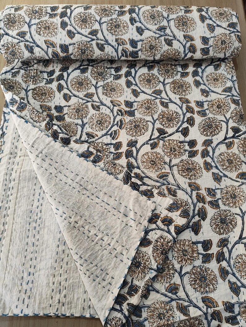 K&ouml;nnte beinhalten: Eine wei&szlig;e und beige gesteppte Decke mit floralem Muster und blauen Akzenten. Die Steppdecke hat ein sich wiederholendes Muster aus gro&szlig;en Blumen und Bl&auml;ttern. Die Kanten sind mit blauem Faden gen&auml;ht. Die Steppdecke ist gefaltet und aufgerollt, wodurch das Muster sichtbar wird.
