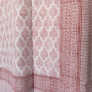 May include: White and red patterned textile. The fabric features a repeating paisley-like design in red, with a border of geometric patterns and vertical stripes. The textile is likely a quilt or blanket.