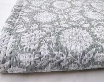 May include: A folded, quilted blanket with a floral pattern in shades of gray and white. The design features large, stylized flowers and leaves, with visible white stitching throughout. The fabric appears soft and textured.