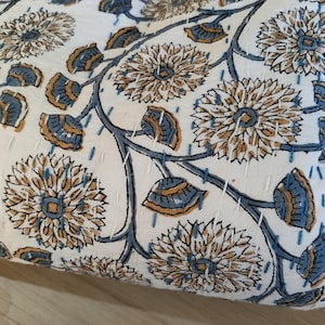 K&ouml;nnte beinhalten: Cremefarbener Stoff mit einem sich wiederholenden Blumenmuster in Beige und Blau. Das Design zeigt gro&szlig;e, stilisierte Blumen und Bl&auml;tterzweige mit sichtbaren wei&szlig;en N&auml;hten. Der Stoff scheint gesteppt zu sein.