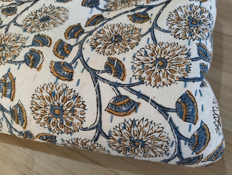 K&ouml;nnte beinhalten: Cremefarbener Stoff mit einem sich wiederholenden Blumenmuster in Beige und Blau. Das Design zeigt gro&szlig;e, stilisierte Blumen und Bl&auml;tterzweige mit sichtbaren wei&szlig;en N&auml;hten. Der Stoff scheint gesteppt zu sein.