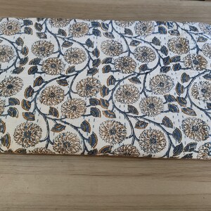 K&ouml;nnte beinhalten: Gefalteter wei&szlig;er Stoff mit einem sich wiederholenden Blumenmuster in Braun- und Blaut&ouml;nen. Das Design zeigt gro&szlig;e, stilisierte Blumen, die durch sich windende Stiele und Bl&auml;tter verbunden sind. Der Stoff ist wahrscheinlich eine Steppdecke oder eine Decke.