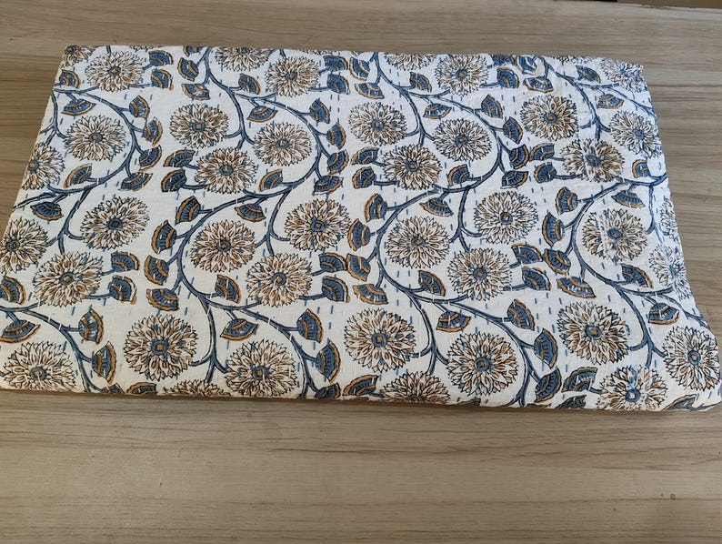 K&ouml;nnte beinhalten: Gefalteter wei&szlig;er Stoff mit einem sich wiederholenden Blumenmuster in Braun- und Blaut&ouml;nen. Das Design zeigt gro&szlig;e, stilisierte Blumen, die durch sich windende Stiele und Bl&auml;tter verbunden sind. Der Stoff ist wahrscheinlich eine Steppdecke oder eine Decke.