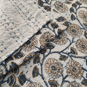 K&ouml;nnte beinhalten: Eine wei&szlig;e Steppdecke mit floralem Muster und blauen und beigen Akzenten. Die Steppdecke hat ein sich wiederholendes Blumendesign mit blauen Stielen und Bl&auml;ttern. Die Steppdecke besteht aus einem weichen, strukturierten Stoff und hat blaue N&auml;hte.