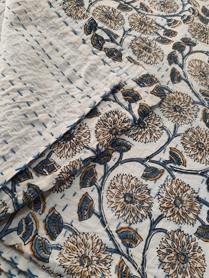 K&ouml;nnte beinhalten: Eine wei&szlig;e Steppdecke mit floralem Muster und blauen und beigen Akzenten. Die Steppdecke hat ein sich wiederholendes Blumendesign mit blauen Stielen und Bl&auml;ttern. Die Steppdecke besteht aus einem weichen, strukturierten Stoff und hat blaue N&auml;hte.