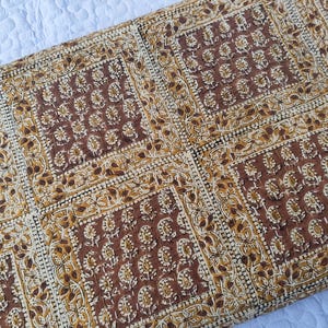 May include: A rectangular, quilted textile with a repeating pattern of brown floral motifs on a cream background. The design features square blocks with detailed floral and leaf patterns, creating a symmetrical and decorative appearance. The fabric appears to be cotton or a similar natural fiber.