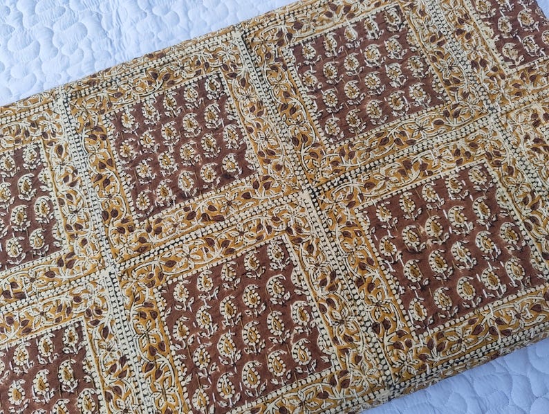 May include: A rectangular, quilted textile with a repeating pattern of brown floral motifs on a cream background. The design features square blocks with detailed floral and leaf patterns, creating a symmetrical and decorative appearance. The fabric appears to be cotton or a similar natural fiber.