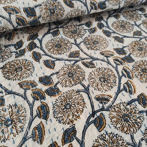 K&ouml;nnte beinhalten: Wei&szlig;er Stoff mit einem sich wiederholenden Blumenmuster in Braun- und Blaut&ouml;nen. Das Design zeigt gro&szlig;e, stilisierte Blumen und Bl&auml;tter, die durch sich windende Stiele verbunden sind. Der Stoff ist mit sichtbaren N&auml;hten gesteppt.