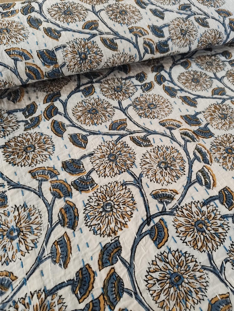 K&ouml;nnte beinhalten: Wei&szlig;er Stoff mit einem sich wiederholenden Blumenmuster in Braun- und Blaut&ouml;nen. Das Design zeigt gro&szlig;e, stilisierte Blumen und Bl&auml;tter, die durch sich windende Stiele verbunden sind. Der Stoff ist mit sichtbaren N&auml;hten gesteppt.