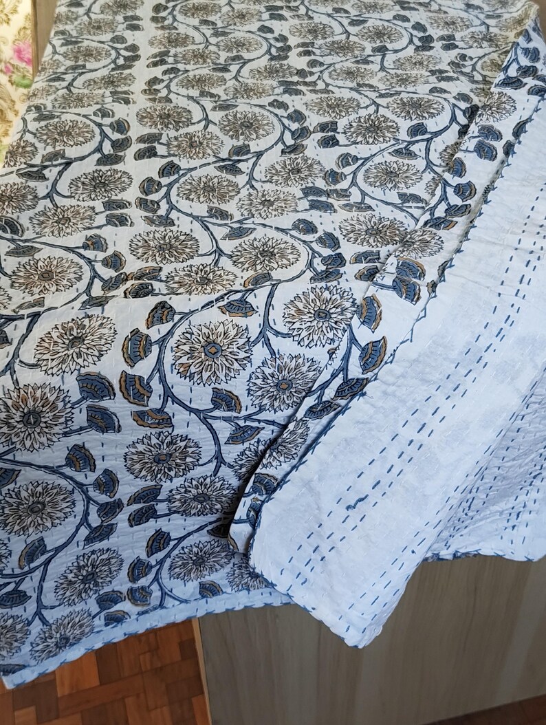 K&ouml;nnte beinhalten: Eine wei&szlig;e und blaue Steppdecke mit Blumenmuster. Die Steppdecke weist ein sich wiederholendes Muster aus gro&szlig;en, stilisierten Blumen und Bl&auml;ttern in Blau-, Braun- und Wei&szlig;t&ouml;nen auf. Die Steppdecke ist gefaltet und zeigt einen wei&szlig;en, gen&auml;hten Rand.