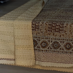 May include: A folded textile with a cream-colored, stitched section and a brown patterned section. The brown section features floral and geometric designs. The cream section has horizontal rows of stitching. The textile appears to be a decorative table runner or throw.