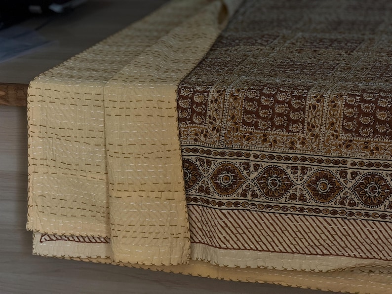 May include: A folded textile with a cream-colored, stitched section and a brown patterned section. The brown section features floral and geometric designs. The cream section has horizontal rows of stitching. The textile appears to be a decorative table runner or throw.