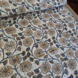 K&ouml;nnte beinhalten: Gerolltes wei&szlig;es Textil mit floralem Muster. Das Design zeigt gro&szlig;e, stilisierte Blumen in Beige- und Blaut&ouml;nen, verbunden durch sich windende Stiele und Bl&auml;tter. Der Stoff scheint gesteppt zu sein, mit sichtbaren N&auml;hten.