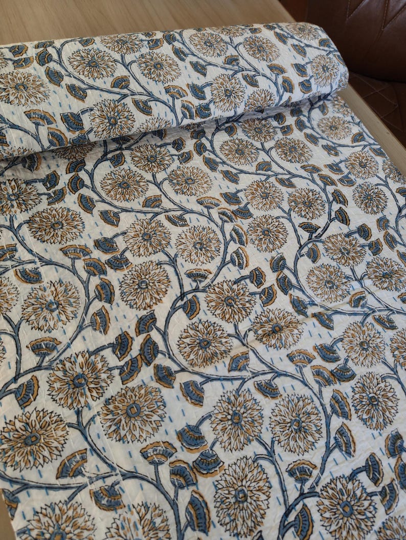 K&ouml;nnte beinhalten: Gerolltes wei&szlig;es Textil mit floralem Muster. Das Design zeigt gro&szlig;e, stilisierte Blumen in Beige- und Blaut&ouml;nen, verbunden durch sich windende Stiele und Bl&auml;tter. Der Stoff scheint gesteppt zu sein, mit sichtbaren N&auml;hten.