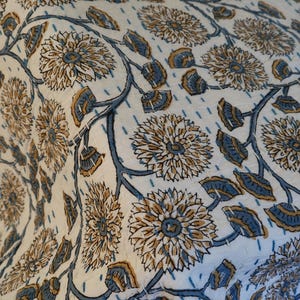 K&ouml;nnte beinhalten: Nahaufnahme eines wei&szlig;en Textils mit einem sich wiederholenden Blumenmuster. Das Design zeigt gro&szlig;e, stilisierte Blumen in Beige- und Blaut&ouml;nen, verbunden durch blaue Stiele und Bl&auml;tter. Der Stoff hat ein gestepptes Aussehen.