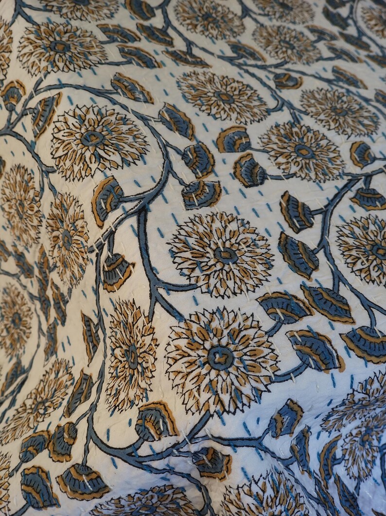 K&ouml;nnte beinhalten: Nahaufnahme eines wei&szlig;en Textils mit einem sich wiederholenden Blumenmuster. Das Design zeigt gro&szlig;e, stilisierte Blumen in Beige- und Blaut&ouml;nen, verbunden durch blaue Stiele und Bl&auml;tter. Der Stoff hat ein gestepptes Aussehen.