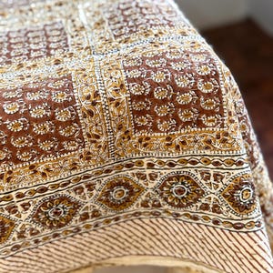 May include: A brown and cream-colored textile with a detailed floral and geometric pattern. The fabric features a repeating design of flowers and shapes in shades of brown, cream, and gold. The edge has a striped pattern.