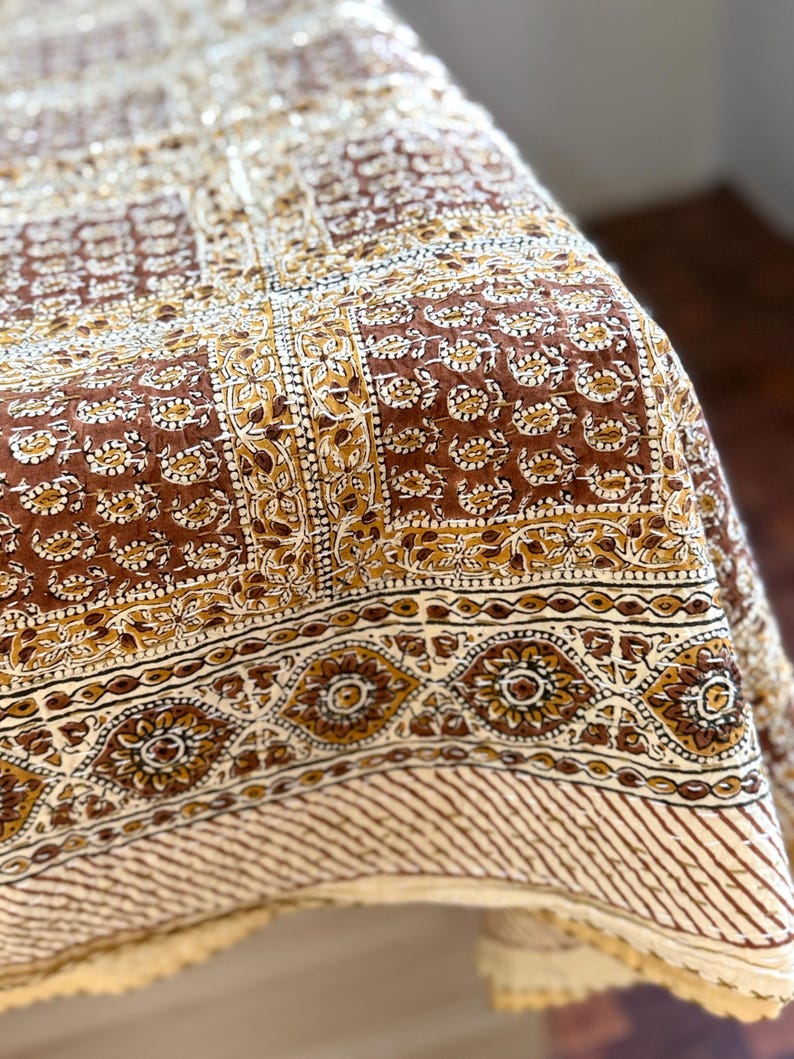 May include: A brown and cream-colored textile with a detailed floral and geometric pattern. The fabric features a repeating design of flowers and shapes in shades of brown, cream, and gold. The edge has a striped pattern.