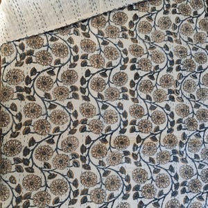 K&ouml;nnte beinhalten: Eine cremefarbene Steppdecke mit einem Blumenmuster in Braun und Marineblau. Die Steppdecke weist ein sich wiederholendes Design aus stilisierten Blumen und Bl&auml;ttern auf. Der obere Rand ist einfarbig cremefarben mit sichtbaren N&auml;hten.