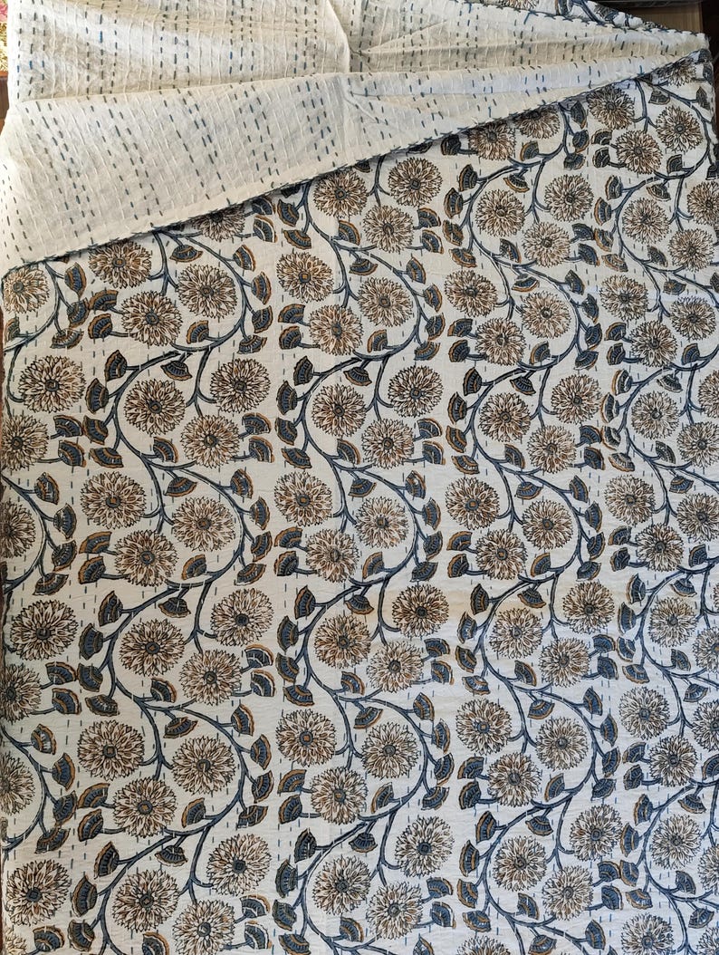 K&ouml;nnte beinhalten: Eine cremefarbene Steppdecke mit einem Blumenmuster in Braun und Marineblau. Die Steppdecke weist ein sich wiederholendes Design aus stilisierten Blumen und Bl&auml;ttern auf. Der obere Rand ist einfarbig cremefarben mit sichtbaren N&auml;hten.