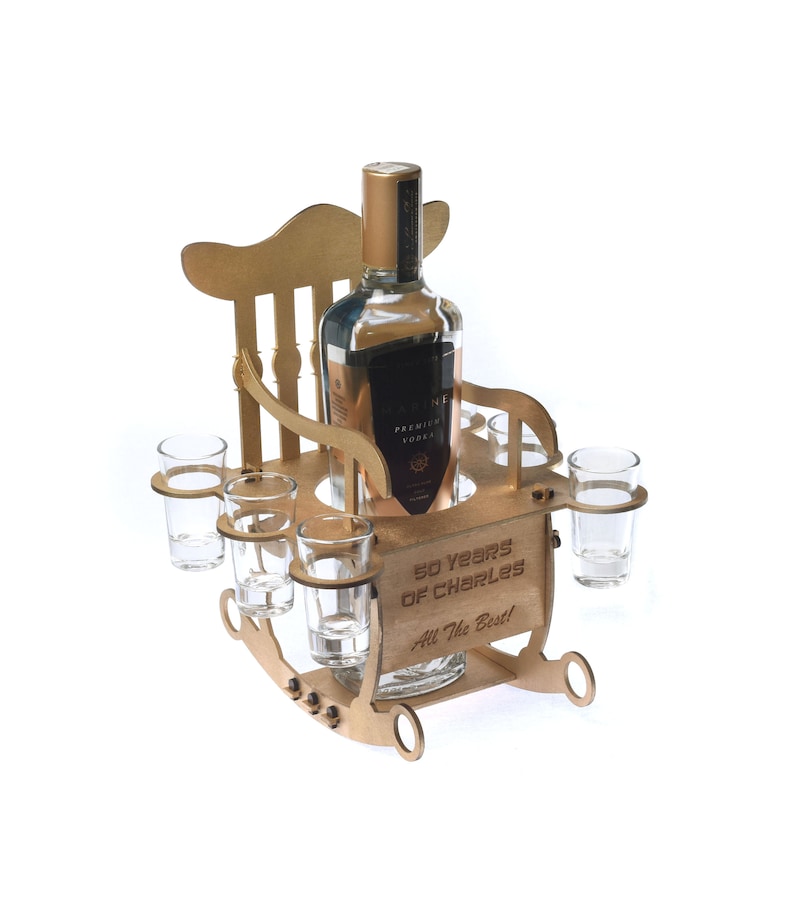 Puede incluir: Un soporte para botellas de madera con forma de mecedora con cuatro vasos de chupito. La silla es dorada y tiene el texto "50 Years of Charles All The Best!" grabado en la parte delantera.