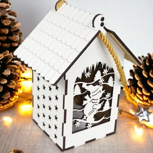 Puede incluir: Adorno navideño de madera blanca con forma de casa con un diseño de muñeco de nieve recortado en la parte delantera. La casa tiene un asa de cuerda y un patrón de estrellas en el lateral.