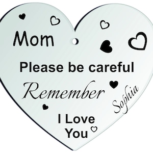Puede incluir: Tres placas blancas en forma de corazón con texto negro. La primera placa dice "Mommy Olivia y Liam conduzcan con cuidado Los esperamos en la cabaña Los queremos". La segunda placa dice "Mamá Por favor, ten cuidado Recuerda a Sophia Te quiero". La tercera placa dice "Eres LA MEJOR MAMÁ DEL MUNDO Amelia Te quiero".