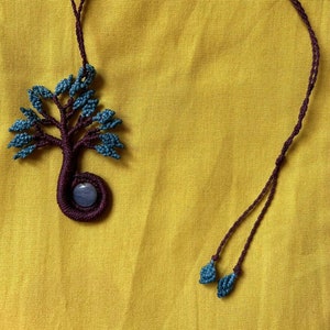 May include: A handmade macrame necklace featuring a tree pendant. The tree has a dark purple trunk and branches with blue leaves. A small, round, blue stone is at the base of the tree. The necklace cord is dark purple.