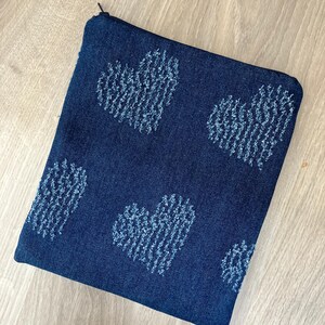 May include: A dark blue denim zippered pouch with a pattern of light blue embroidered hearts. The rectangular pouch is on a light wood surface. The zipper is black.