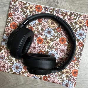 Peut inclure: Casque audio sans fil noir avec un étui de transport à motif floral. L'étui est doté d'une fermeture à glissière et d'un fond blanc avec des fleurs roses, orange et bleues.