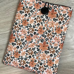 Puede incluir: Una funda de libro rectangular con estampado floral y cierre de botón negro. La funda presenta un patrón repetido de flores naranjas y marrones con hojas verdes sobre un fondo blanco. Los bordes son negros.