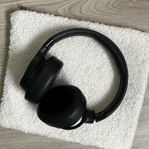Könnte beinhalten: Schwarze kabellose Kopfhörer mit einer weichen, weißen, flauschigen Tasche. Die Kopfhörer haben einen schwarzen Kopfbügel und Ohrmuscheln.