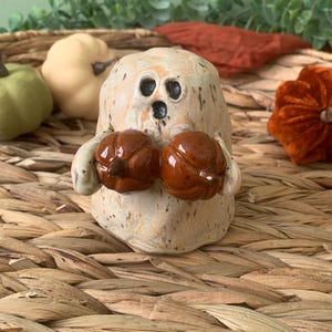 May include: A ceramic ghost figurine with a white and brown speckled glaze. The ghost is holding two small, brown pumpkins in its hands.