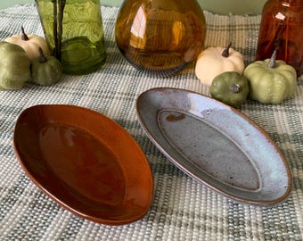May include: Two oval ceramic serving dishes, one brown and one blue-grey, on a white and grey checkered surface.