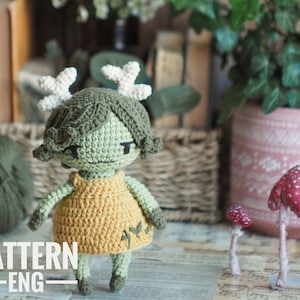 May include: Crochet pattern for a green amigurumi doll with antlers, wearing a yellow dress and standing on a table with a music sheet and mushrooms.
