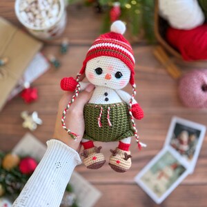 K&ouml;nnte beinhalten: Geh&auml;kelte Schneemann-Puppe mit rot-wei&szlig; gestreifter M&uuml;tze, roten F&auml;ustlingen und gr&uuml;nem Rock. Der Schneemann hat eine Karottennase und Knopfaugen. Er tr&auml;gt braune Rentierschuhe. Eine festliche Weihnachtsdekoration.