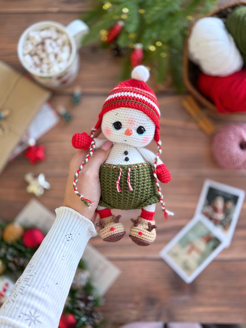 K&ouml;nnte beinhalten: Geh&auml;kelte Schneemann-Puppe mit rot-wei&szlig; gestreifter M&uuml;tze, roten F&auml;ustlingen und gr&uuml;nem Rock. Der Schneemann hat eine Karottennase und Knopfaugen. Er tr&auml;gt braune Rentierschuhe. Eine festliche Weihnachtsdekoration.