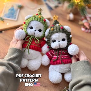 Puede incluir: Dos osos de peluche blancos de crochet que llevan gorros de árbol de Navidad verdes y rojos. Los osos sostienen un cartel que dice "PDF CROCHET PATTERN ENG" con una bandera estadounidense.