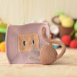 Puede incluir: Un juguete de crochet con un cuerpo morado, una cola marr&oacute;n y una cara marr&oacute;n con dos ojos. El juguete est&aacute; sentado sobre una superficie marr&oacute;n claro.